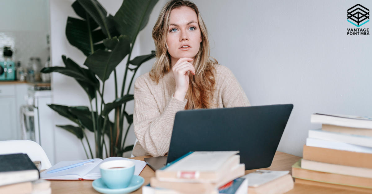 A woman sitting at a table with a laptop, surrounded by books and a cup of coffee, looking thoughtful as she works.