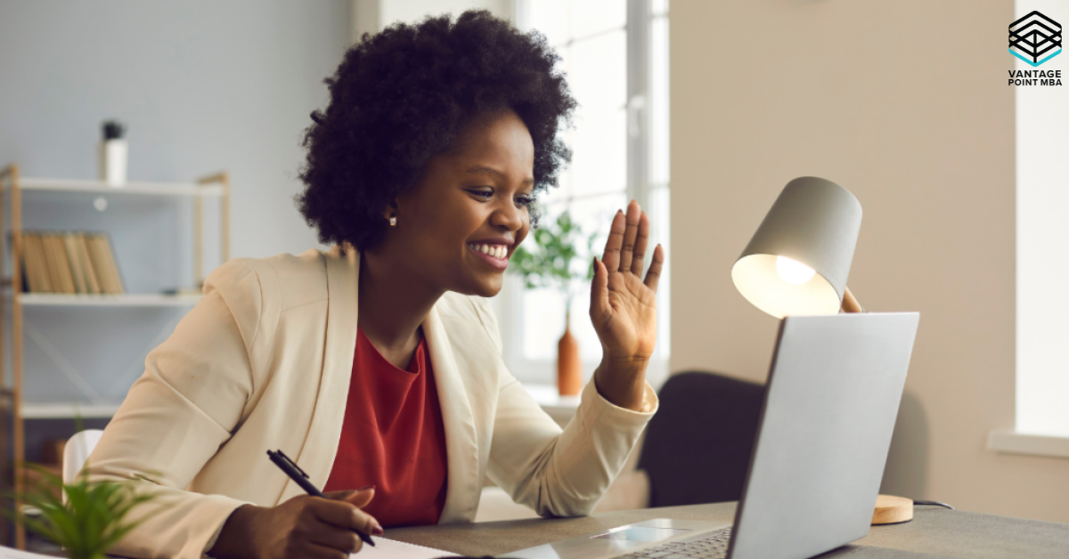 A smiling woman sits at a desk in a well-lit home office, waving during a video call on her laptop while holding a pen and notebook, representing confident preparation for a virtual Wharton MBA interview.